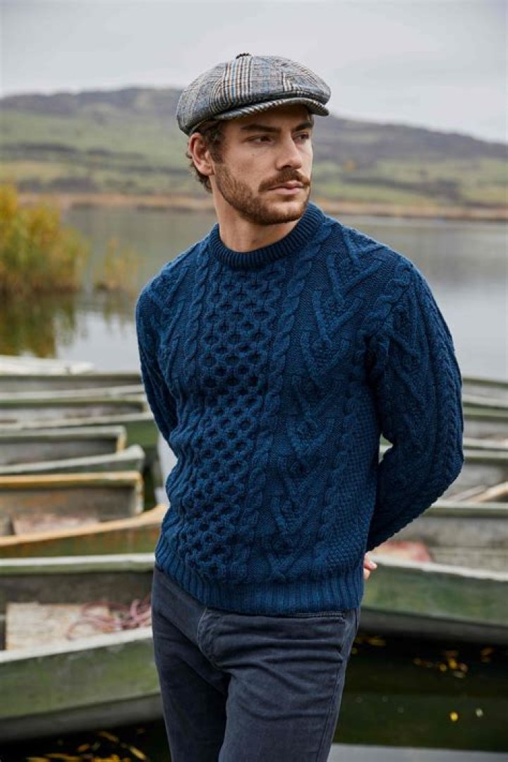 A man in a knit reindeer Christmas cardigan button up sweater, complete with matching red bow tie and a classy mustache. He straightens his bow tie with a cheesy smile on his face, proud of his fashion style. Damask style vintage wall paper in the background. Horizontal with copy space | Image: Unsplash