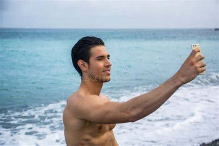 a shirtless young man taking a selfie in front of a blue wall with his